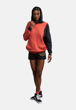 Hoodie in koraal en zwart, met een losse pasvorm en textiel stof. Gecombineerd met zwarte shorts en hoge sneakers.