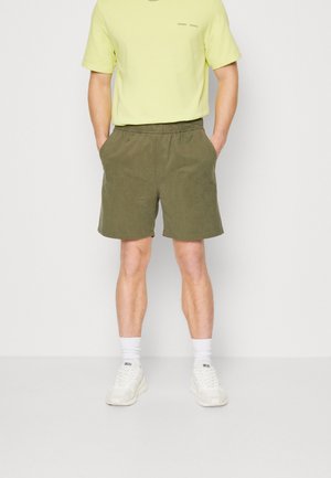 Homme portant une chemise à manches courtes vert clair, un short à taille élastique vert olive, des chaussettes blanches et des baskets blanches Diesel, debout les mains dans les poches.