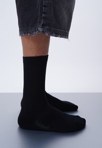 Schwarze gerippte Knöchelsocken aus einem weichen Material mit nahtlosem Zehenbereich und eingesticktem Logo nahe der Ferse, kombiniert mit ausgefranstem schwarzen Denimshorts.