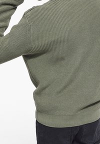 Maglione a collo alto di colore verde oliva, con un motivo a trama, fondo a coste e accenti sulle spalle. Il tessuto appare morbido, con maniche lunghe e vestibilità ampia.