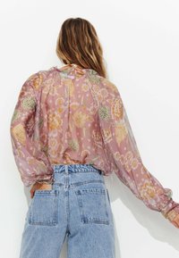 Blouse transparente à fleurs de couleur rose clair avec des manches bouffantes. Présente un décolleté froncé et des motifs floraux complexes. Associée à un jean taille haute.