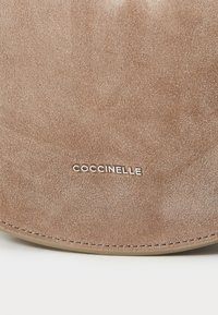 Borsa in suede marrone con una superficie liscia, caratterizzata da un bordo cucito e un logo argentato. Il design è arrotondato con una leggera lucentezza.