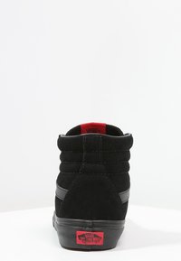 Sneaker alta nera in suede con colletto imbottito, dettagli rossi e suola in gomma che presenta il logo "VANS OFF THE WALL" sul tallone.