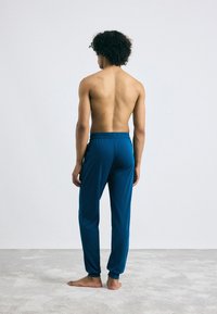 Blauwe loungebroek met een elastische tailleband, tapered benen en een gladde textuur, te zien aan een persoon met blote huid van achteren.
