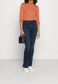 Höga flarejeans i mörkblå denim, kombinerat med en orange långärmad polotröja och vita sneakers, hållande en svart handväska.