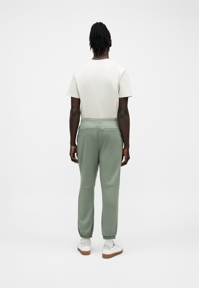 Personne debout de dos, portant une chemise blanche à manches courtes, un pantalon de jogging vert, des chaussettes blanches et des baskets blanches sur un fond blanc.