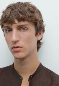 Male model with short, wavy brown hair, wearing a dark brown button-up shirt. Clear skin and neutral background.