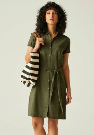 Femme aux cheveux bouclés portant une robe verte boutonnée nouée à la taille, portant un sac à bandoulière rayé noir et blanc avec des bretelles marron.