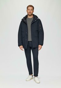 Parka marine avec une capuche, dotée de poches frontales et d'une fermeture éclair. Portée sur un pull gris, accompagnée d'un pantalon marine et de baskets blanches.