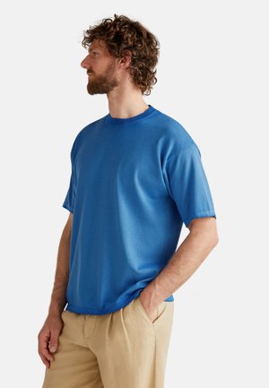 Camiseta de cuello redondo de manga corta en azul, hecha de tejido suave, con un corte relajado y diseño en un solo color. Llevada con pantalones de color claro.