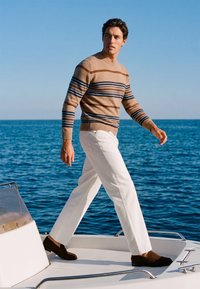 Homme portant un pull beige rayé et un pantalon blanc marchant sur le pont d'un bateau avec la mer et un ciel dégagé en arrière-plan.