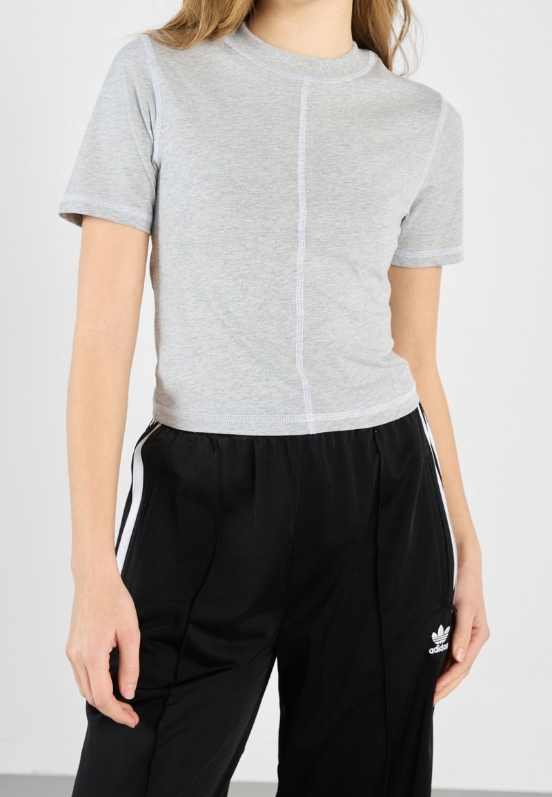 Jeune personne portant un t-shirt gris clair ajusté à manches courtes et un pantalon de survêtement Adidas noir ample avec des bandes blanches.