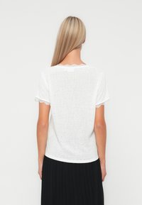 Hvid kortærmet T-shirt med blondertrim ved ærmerne og halsudskæringen, lavet af letvægtsstof, vist mod en ensfarvet baggrund.
