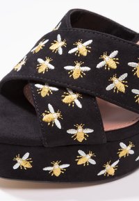 Sandale plateforme en daim noir avec des motifs de guêpe brodés en or et blanc couvrant les brides et le bord de la semelle.