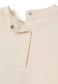 Encolure de vêtement beige avec un col côtelé et deux boutons-pression en métal partiellement défaits sur fond blanc.