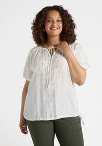 Blusa bianca con maniche corte, tessuto strutturato, punti ricamati in oro e scollatura con cordoncino. Abbinata a pantaloni verdi.