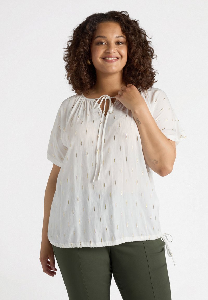 Blusa bianca con maniche corte, tessuto strutturato, punti ricamati in oro e scollatura con cordoncino. Abbinata a pantaloni verdi.