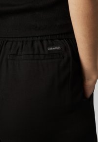 Nærbillede af en person iført sorte Calvin Klein-bukser med elastisk talje og en baglomme, kombineret med en sort ribbet top.