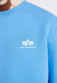 Sudadera azul con un logo blanco de Alpha Industries en el pecho, mostrando parte del cuello y el área del hombro.