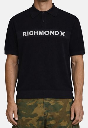 Uomo con polo nera a maniche corte con la scritta "RICHMOND X" sul petto e pantaloni mimetici verdi.