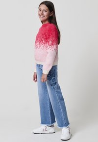 Une fille souriante se tient avec un pull en laine floue rose-rouge, un jean large brodé bleu et des baskets blanches devant un fond clair.
