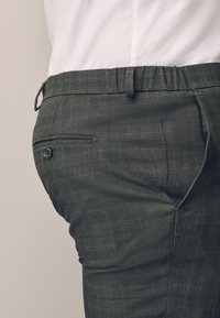 Close-up of a person wearing dark gray plaid dress pants with a back buttoned pocket and a white shirt tucked in.