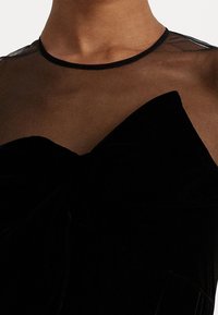 Svart sammetklänning med en genomskinlig meshkrage, med en framträdande rosettdetalj framtill och en mjuk, texturerad tyg.