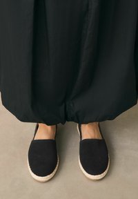 Musta mokkanahkainen espadrille, jossa on pyöreä kärki, juuttikuituvälikerros ja joustavat sivut, yhdistettynä ilmavaan mustaan vaatteeseen.