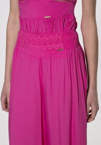 Jumpsuit senza maniche rosa con vita arricciata, dettagli in pizzo e accenti dorati lucidi vicino alla vita. Tessuto morbido e fluido.