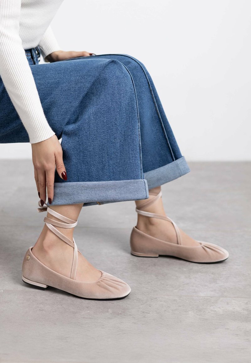 Bailarinas de ante beige con diseño plisado, cintas entrelazadas alrededor del tobillo, combinadas con una falda de mezclilla con dobladillo. Textura suave, perfil bajo.