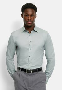 Hellgrünes Hemd mit strukturiertem Kragen, langen Ärmeln und dunklen Knöpfen, kombiniert mit grauen Hosen und einem schwarzen Gürtel.