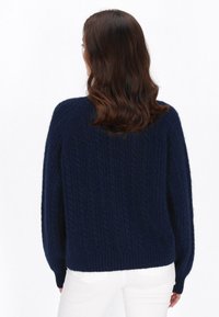 Maglione a maglia cable blu navy con maniche larghe e orlo a coste, realizzato in un tessuto morbido e strutturato. I pantaloni bianchi sono parzialmente visibili.
