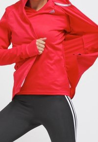 Rød sportsjakke med lynlåsåbning foran, lange ærmer og tommelfingerhuller, lagt over en matchende skjorte. Sorte leggings med hvide striber.