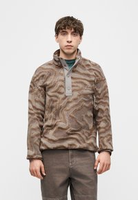 Νεαρός άνδρας που φοράει καφέ φλις πουλόβερ με σχέδιο, με κουμπιά τύπου σναπ και κάθετρη τσέπη στο στήθος, στέκεται μπροστά σε απλό λευκό φόντο.