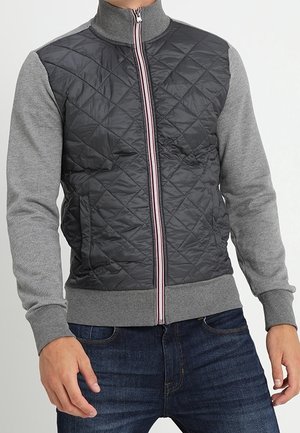 Mann trägt eine gesteppte schwarz-graue Zip-Up-Jacke mit grauen Ärmeln und blauen Jeans und steht mit den Händen in Hüfthöhe.
