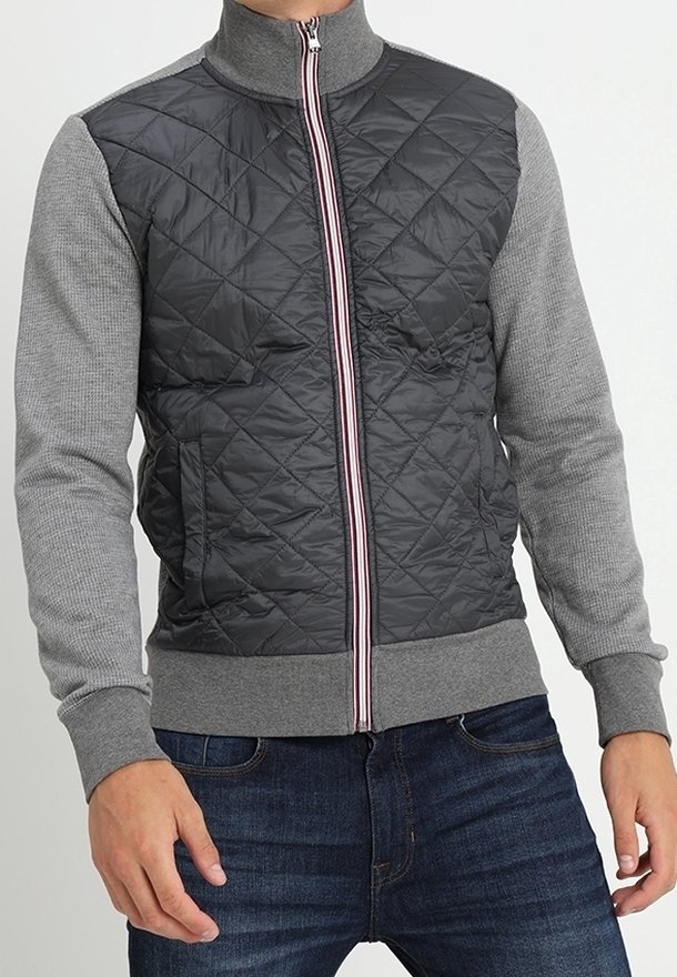 Homme portant une veste matelassée noire et grise zippée avec des manches grises et un jean bleu, debout, mains proches des hanches.