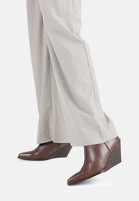 Botines de cuña de cuero marrón con punta afilada y acabado texturizado, combinados con pantalones de pierna ancha en gris claro.