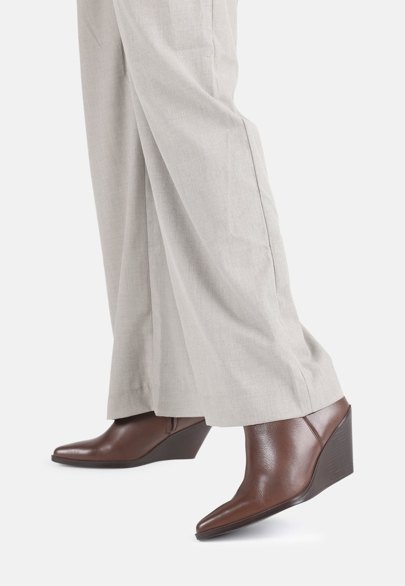 Botines de cuña de cuero marrón con punta afilada y acabado texturizado, combinados con pantalones de pierna ancha en gris claro.