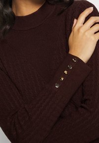Maglione a collo alto in maglia bordeaux con una texture a coste, caratterizzato da accenti in bottone di ottone lungo le maniche.