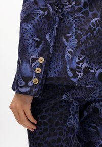 Blaue Stoff mit Leopardenmustern, versehen mit goldenen Knopfakzenten an den Ärmeln. Glatte Textur, taillierter Schnitt.