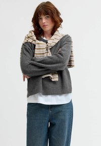 Grå stickad tröja med vida ärmar, i kombination med en vit skjorta och en beige randig sjal hängande runt axlarna. Blå jeans synliga.