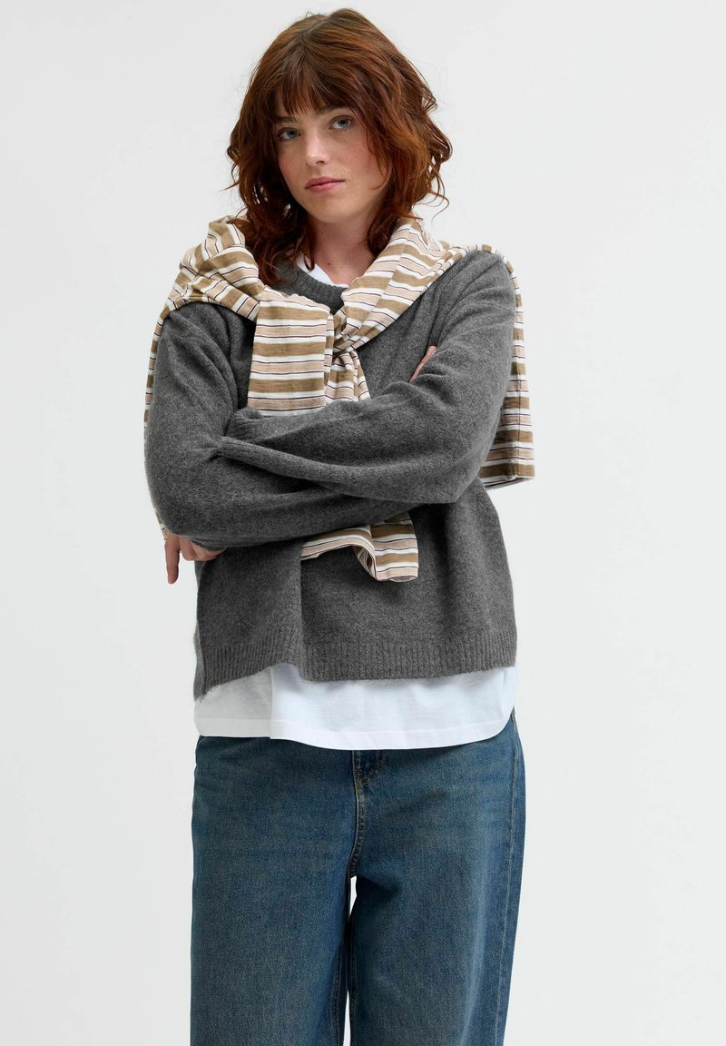 Grå stickad tröja med vida ärmar, i kombination med en vit skjorta och en beige randig sjal hängande runt axlarna. Blå jeans synliga.