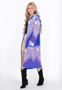 Long cardigan bleu en tricot avec des motifs géométriques en rouge et blanc. Il a une ouverture à l'avant et des manches longues, fabriqué en matériau doux et duveteux.