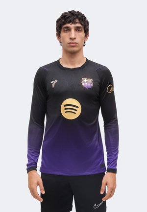 Joven de pie de frente con camiseta negra y morada del FC Barcelona con el logo de Spotify, el escudo del equipo y la marca Nike, manos a los lados.