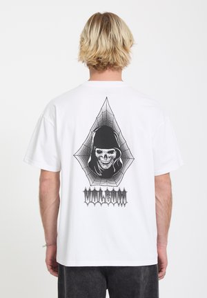 Homme aux cheveux blonds portant un t-shirt blanc avec un motif de squelette noir en capuche et une toile d'araignée, debout devant un fond uni.