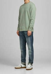 Jasnozielona bluza z małymi białymi zdobieniami, zestawiona z niebieskimi dżinsami i białymi trampkami z brązowymi podeszwami oraz czarnymi akcentami.