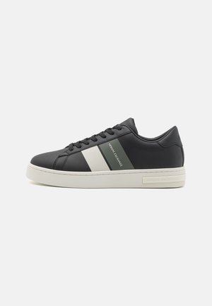 Sneaker in pelle nera con accenti a strisce bianche e verdi, lacci piatti e suola in gomma bianca testurizzata con branding sul lato.