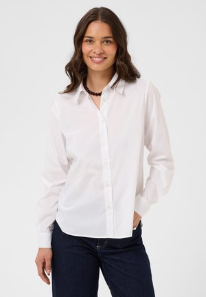 Femme aux cheveux bruns ondulés souriante, portant une chemise blanche boutonnée, un jean sombre et un collier de perles foncé, debout devant un fond uni.