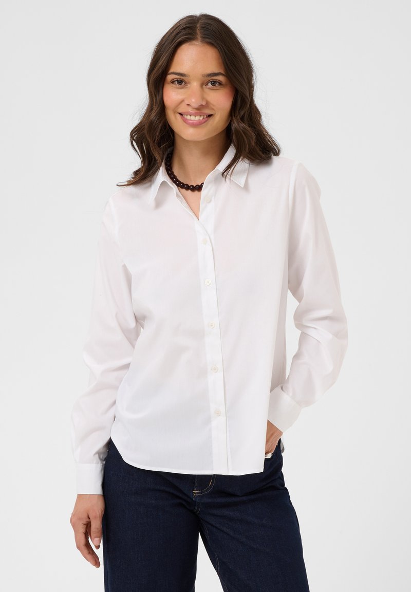 Femme aux cheveux bruns ondulés souriante, portant une chemise blanche boutonnée, un jean sombre et un collier de perles foncé, debout devant un fond uni.