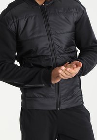 Veste noire matelassée avec fermeture éclair et panneaux en tissu contrasté, dotée de manches longues et d'une coupe ajustée.